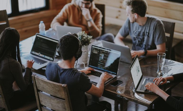 Image shows a technology business workplace with a group of people gathered around a table with laptops