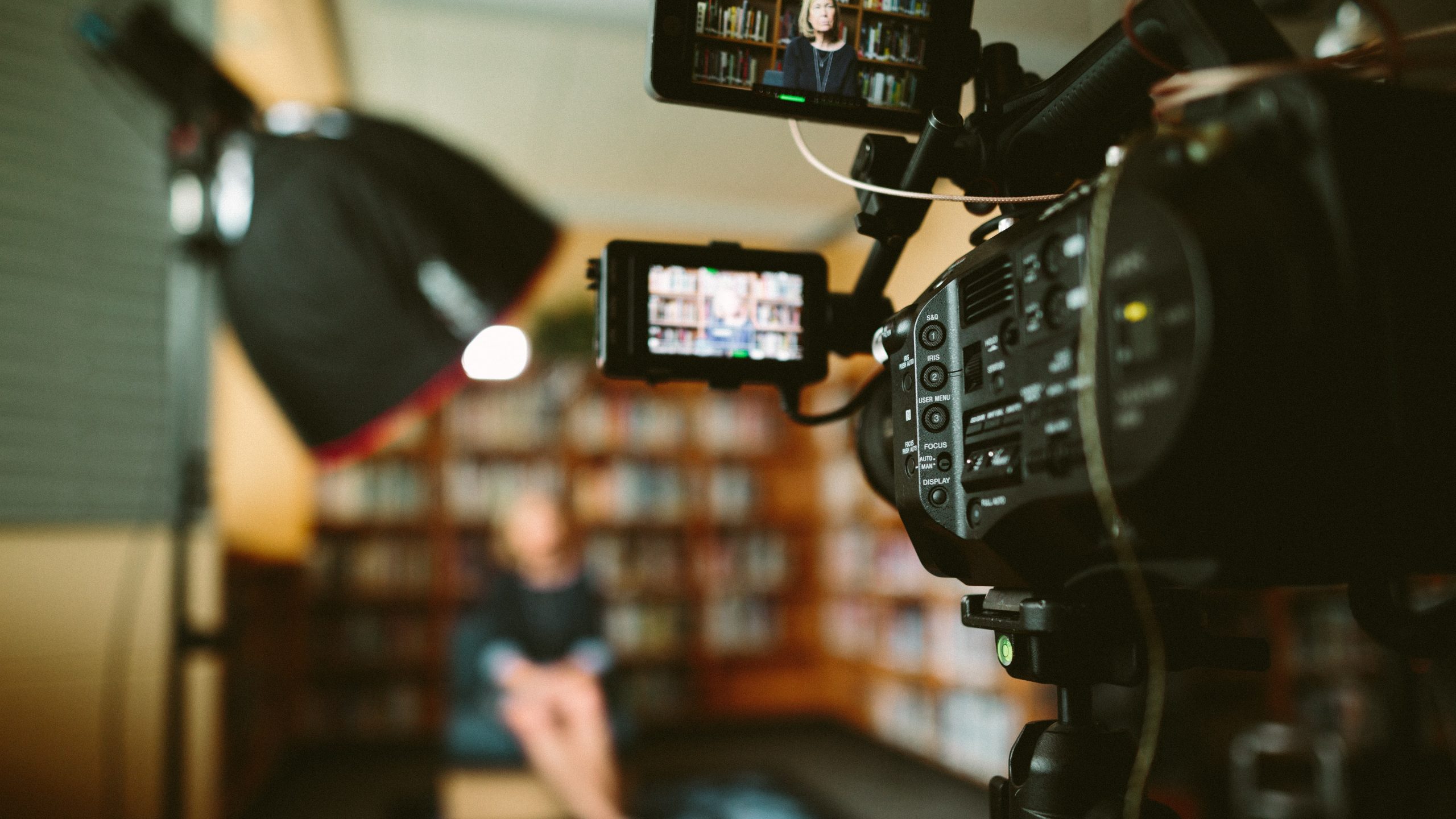 Image shows a camera filming a sit-down video of someone surrounded by lighting and bookshelves