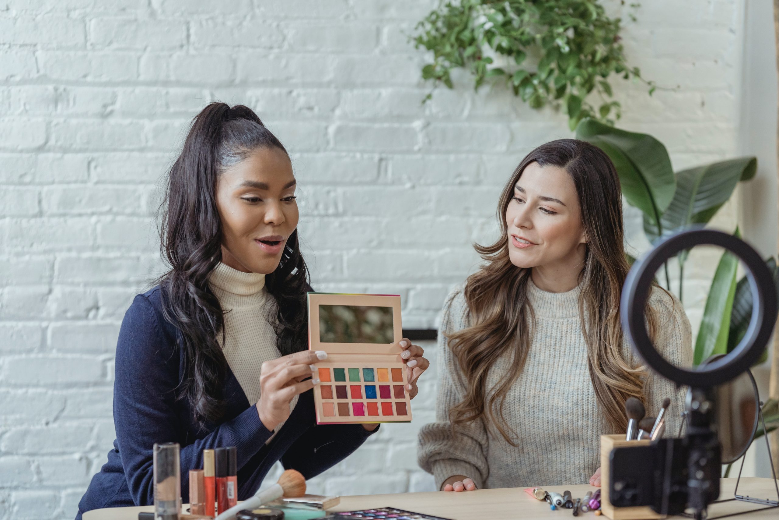 Image shows two women in front of a ring light and camera talking about make up products