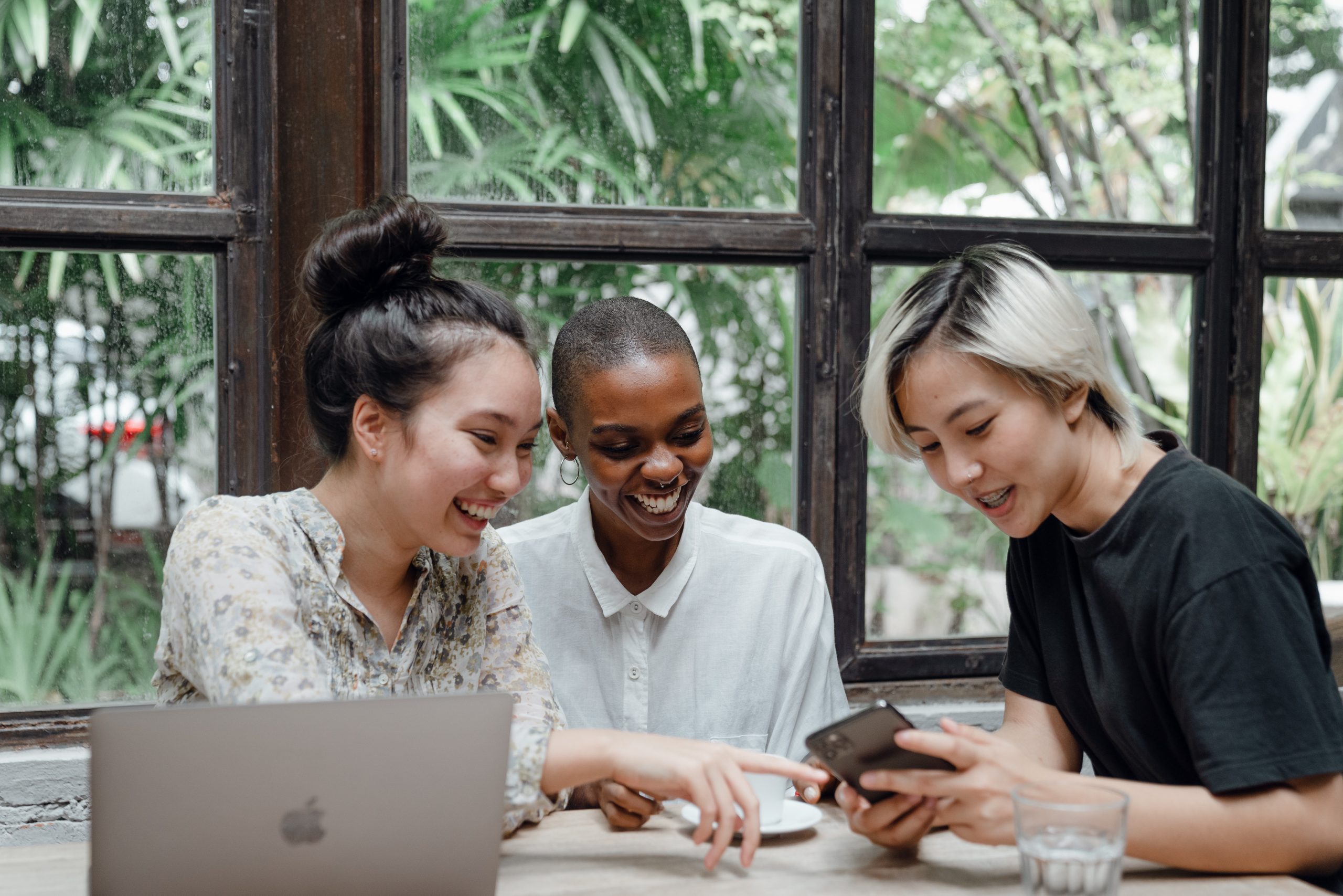 Image shows three young women sat around a laptop and phone. They are watching a crowdfunding video on the phone.