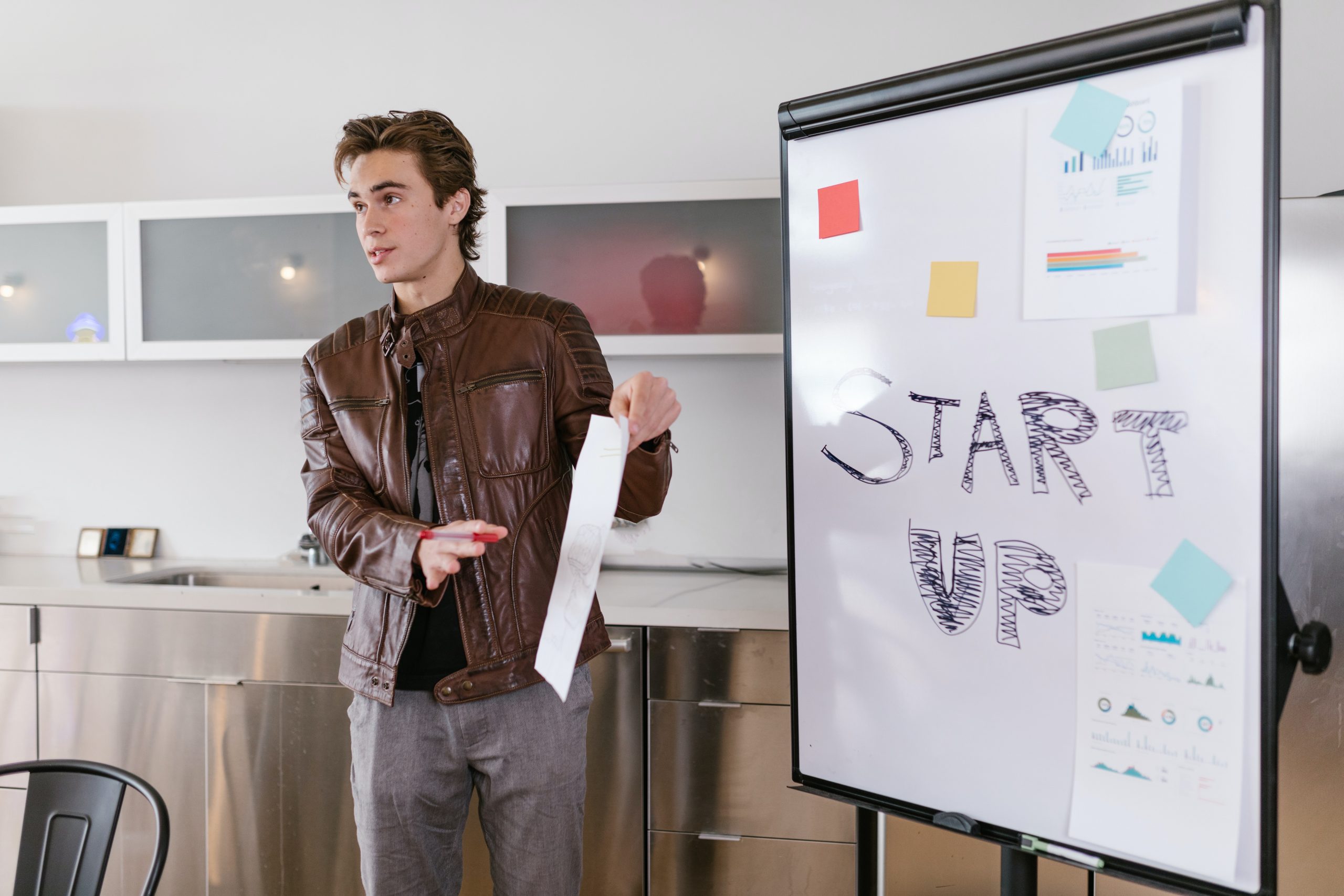 Image shows a young man at a white board holding some paper and explaining his crowdfunding project to an audience. The whiteboard reads "Start Up".