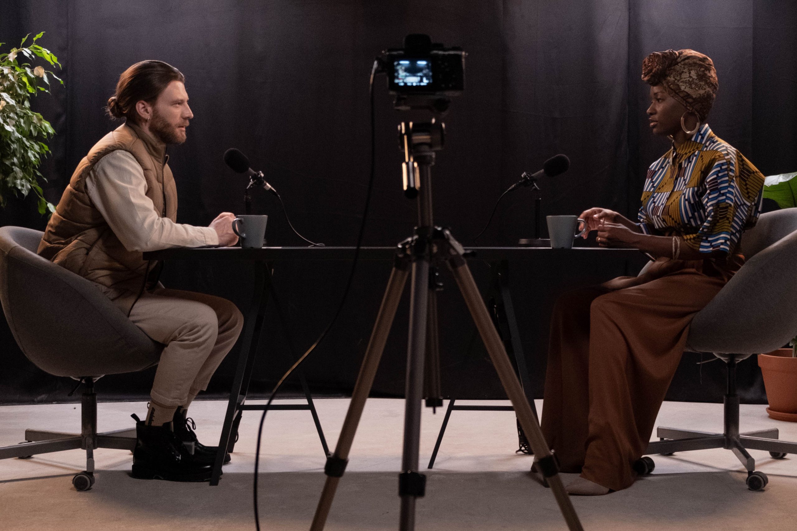 Image shows a man and a woman recording a video podcast. You can see them both sat at a desk with a microphone each and a camera set up in between them.