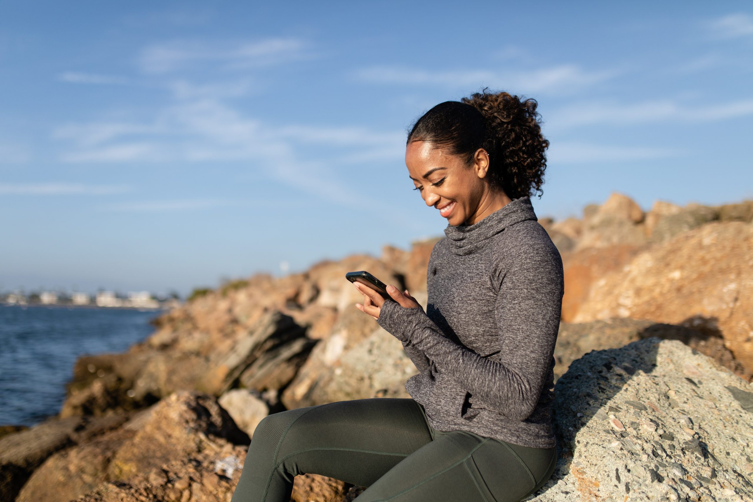 Image shows a woman sat on some rocks by the peach watching video on her mobile phone