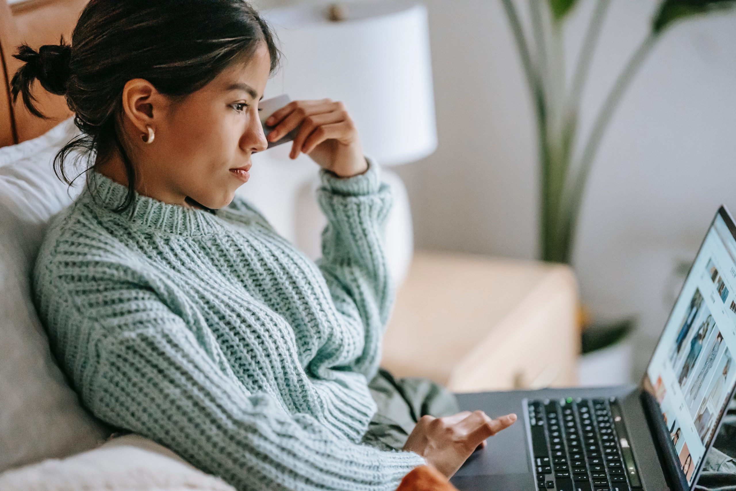 Image shows a woman looking at products on a laptop, holding a bank card in her hand. She looks like she's considering her purchase.