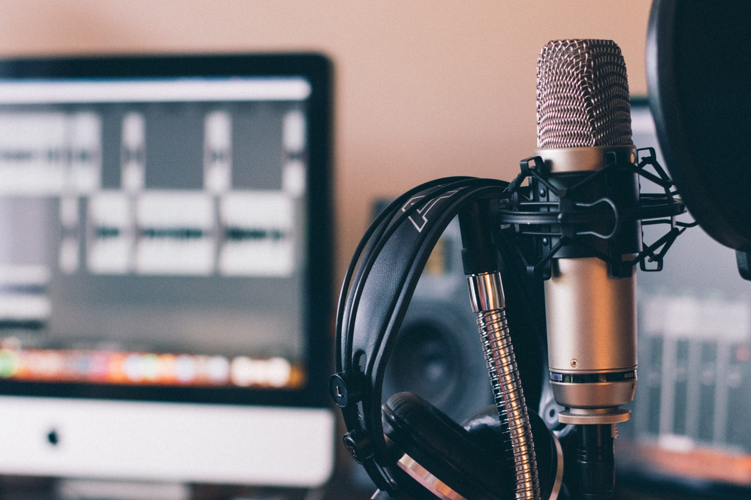 Image shows a microphone with some headphones hung up next to it in the foreground. In the background is a computer screen on which a video podcast is being edited.